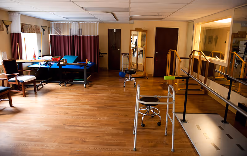 A rehabilitation room with wooden flooring featuring parallel bars for walking exercises, a walker, stools, chairs, a therapy table with equipment, and a large mirror on the wall. The room has beige walls and ceiling tiles with fluorescent lighting.