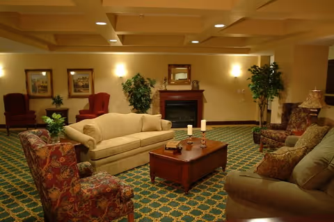 A cozy living room area with patterned carpet, beige and floral upholstered sofas and armchairs arranged around a wooden coffee table with candles and books. The room features a fireplace with a mirror above it, wall sconces providing warm lighting, framed artwork on the walls, and potted plants adding greenery.