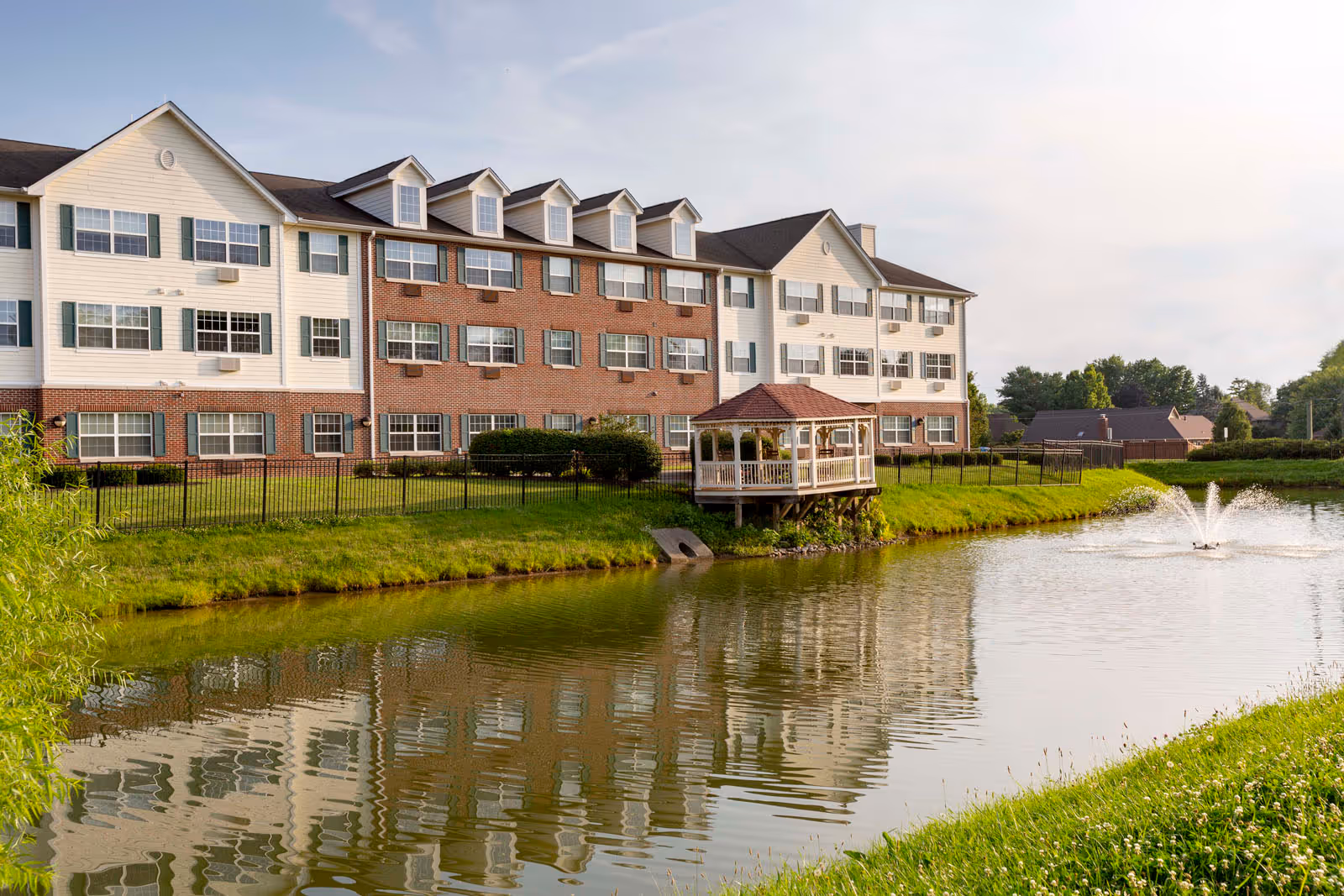 Three-story brick-and-siding senior living building beside a pond with a gazebo and fountain.