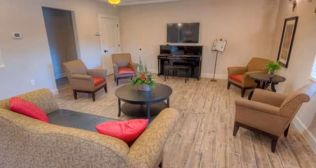 A cozy living room with a patterned sofa adorned with red pillows, four armchairs with red cushions, a round black coffee table with a floral arrangement, a piano with a TV mounted above it, and a music stand in the corner. The room has wood-look tile flooring and neutral-colored walls.