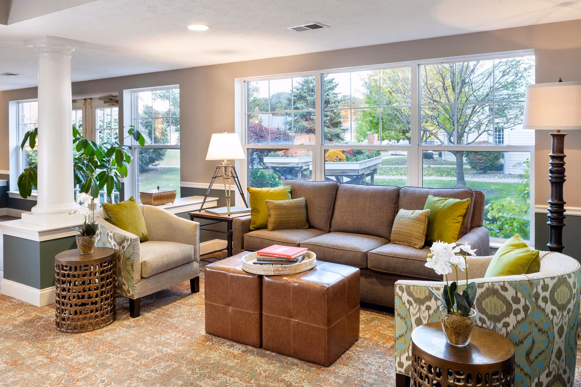 A bright and cozy living room area with a large window showing a garden outside. The room features a brown sofa with green and brown cushions, two armchairs with green pillows, a pair of brown leather ottomans serving as a coffee table with books on top, two side tables with lamps, and decorative plants. The walls are painted beige with white trim and a column is visible on the left side.