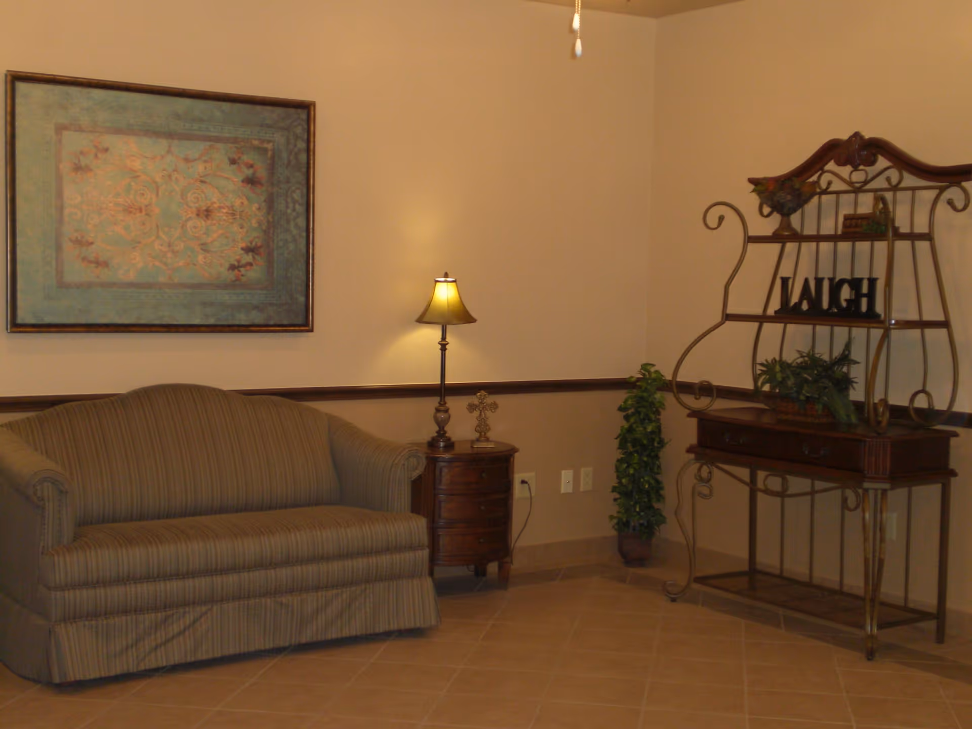 A cozy sitting area with a striped sofa, a small wooden side table with a lamp and decorative cross, a potted plant, and a decorative metal shelf with the word 'LAUGH' displayed on it. A framed artwork hangs on the wall above the sofa.