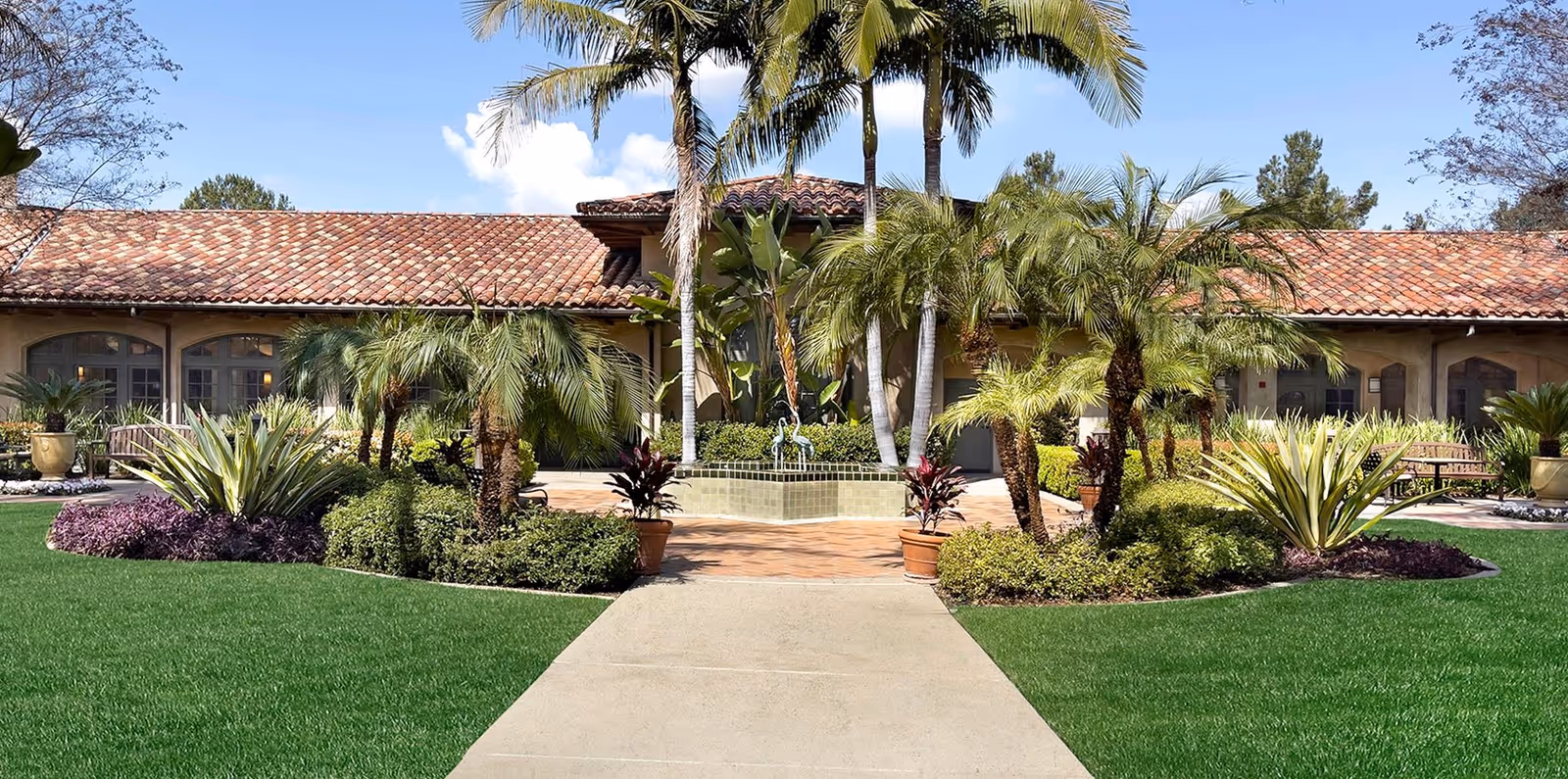 Well-manicured courtyard with palm trees and a central fountain in front of a tile-roofed building.