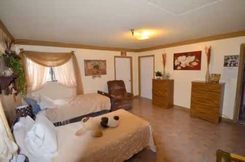 A cozy bedroom in a senior living facility with two beds covered in beige bedspreads, a brown recliner chair, two wooden dressers, wall decorations including a floral painting and a framed picture, and a window with sheer curtains allowing natural light.