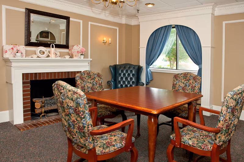 A cozy room with a wooden square table surrounded by four floral upholstered chairs and one dark green leather armchair. There is a white fireplace with decorative plates and a mirror above it. A window with blue curtains lets in natural light, and the walls are painted beige with white trim.