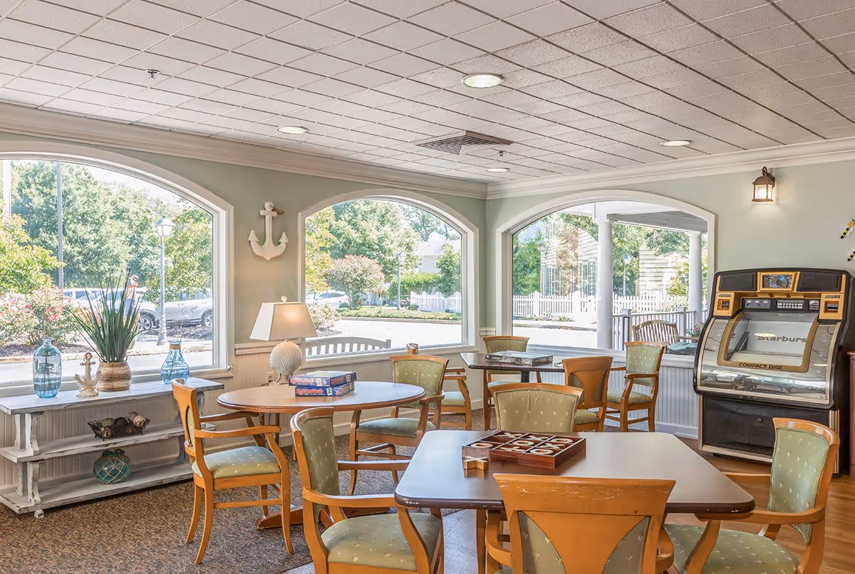 A bright and cozy common area with large arched windows letting in natural light. The room contains several wooden tables and chairs with green cushioned seats. On one table, there are board games, and a vintage jukebox is visible in the corner. Decorative items like vases and a lamp are placed on a white shelf by the windows, and an anchor wall decoration hangs on the wall.