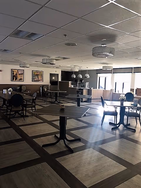 A spacious dining room with multiple tables and chairs arranged neatly. The room has a patterned floor with light and dark wood tones, several ceiling light fixtures, and large windows allowing natural light to enter. There are framed pictures on the far wall and a counter area in the background.