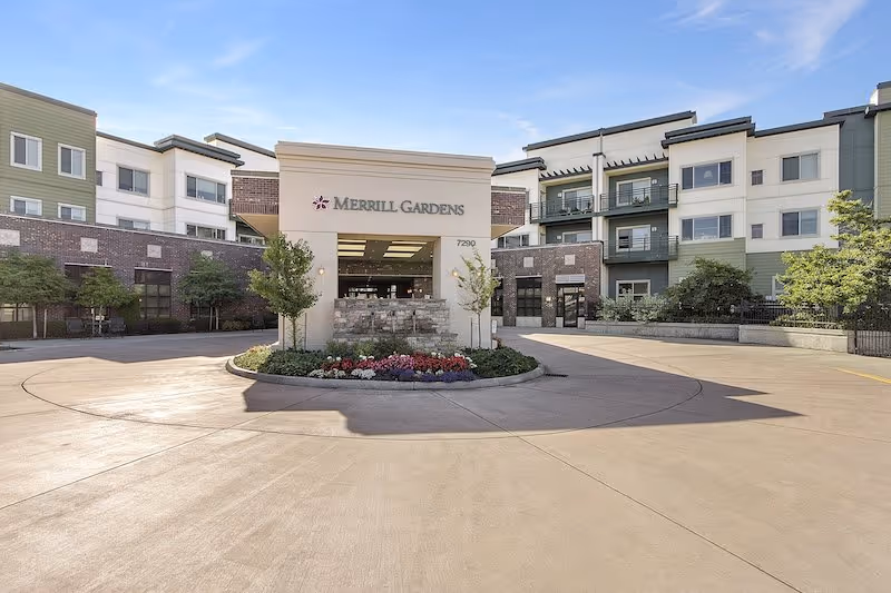 Front exterior view of Merrill Gardens senior living facility with a circular driveway, landscaped flower bed, and multi-story building under a clear blue sky.