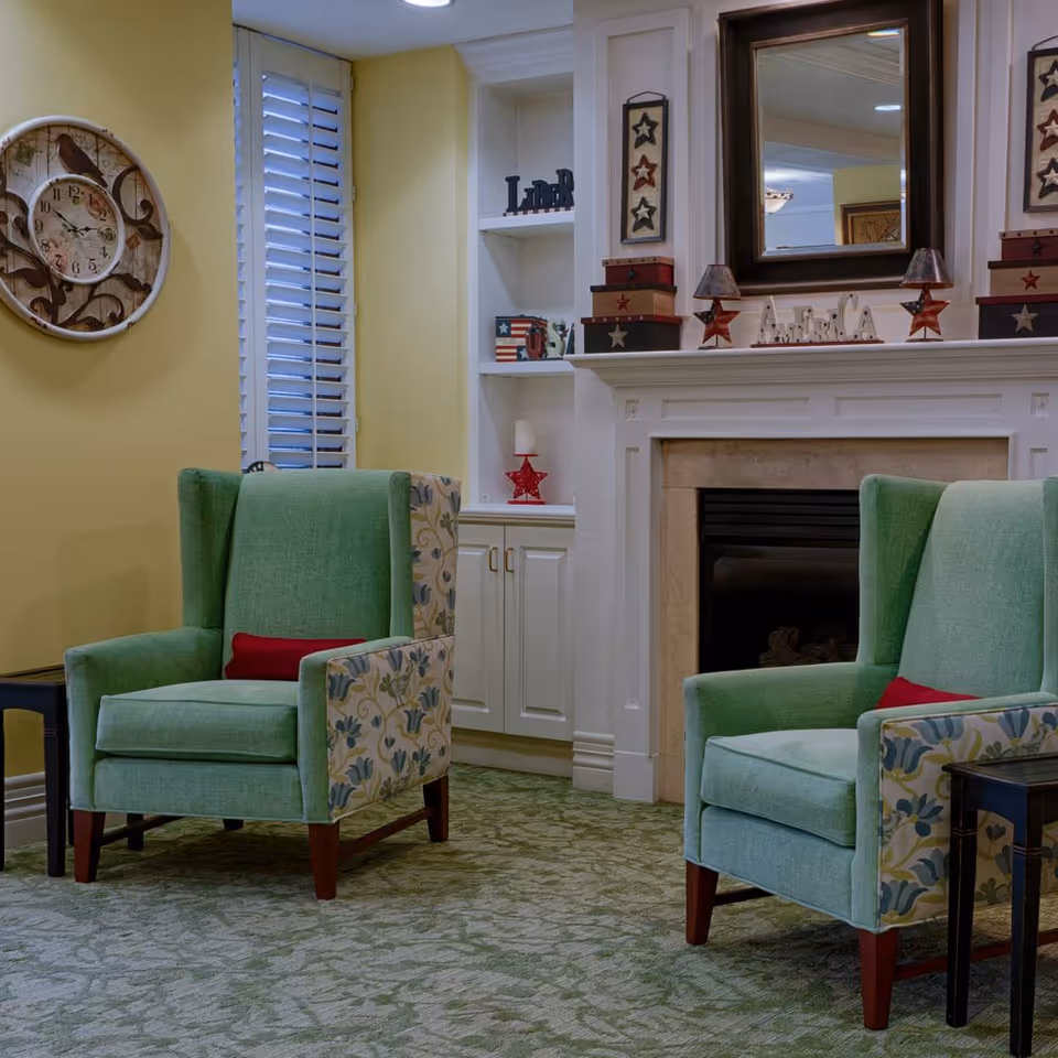 A cozy living room area with two green upholstered armchairs featuring floral patterned sides and red cushions. The room has a green patterned carpet, a white fireplace with decorative items including star-themed decorations and a mirror above it. There is a built-in white shelving unit with various decorative items and a large round wall clock on a yellow wall next to a window with white shutters.
