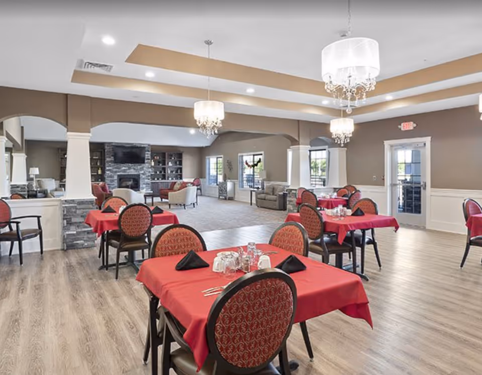 A spacious dining area with multiple tables covered in red tablecloths and set with black napkins, glasses, and silverware. The room features wood flooring, beige walls, and elegant chandeliers hanging from a tray ceiling. In the background, there is a cozy sitting area with armchairs and sofas arranged around a stone fireplace with a mounted TV above it. Large windows allow natural light to enter the room.