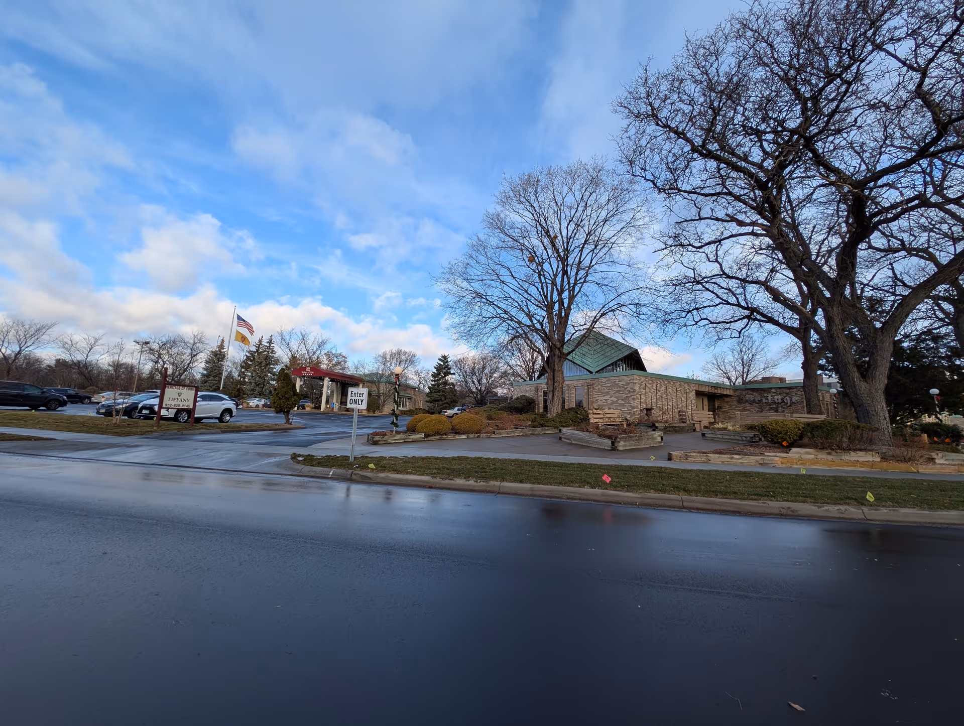 Exterior view of Heritage Of Edina II facility showing a brick building with a green roof, large leafless trees, a parking lot with several cars, and a sign near the entrance. The sky is partly cloudy with blue patches.