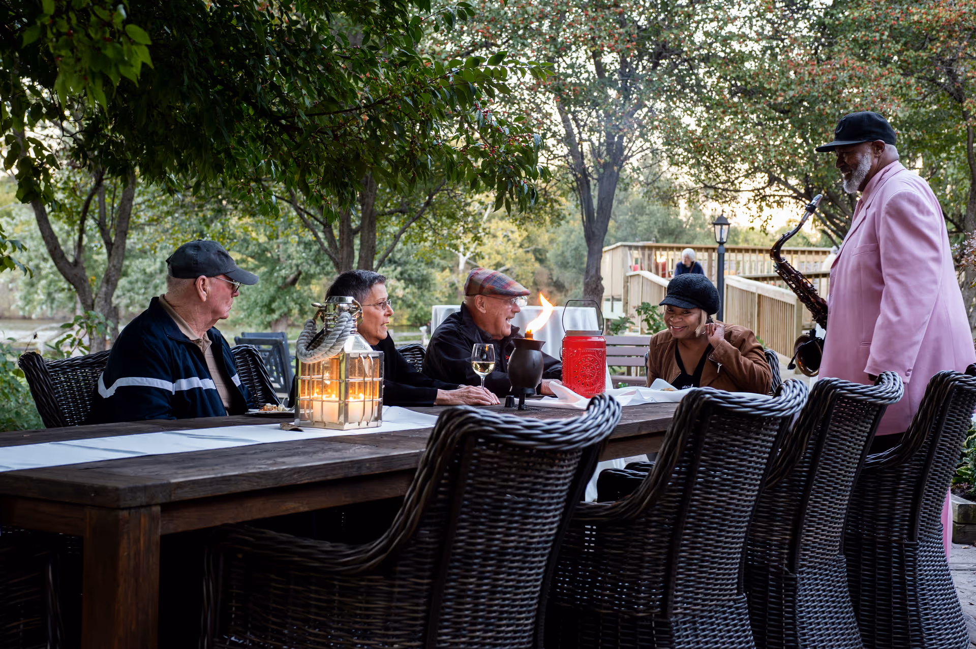 A group of elderly people sitting around a wooden outdoor table under trees, enjoying a social gathering with candles and a tiki torch on the table. A man in a pink suit and black hat is playing a saxophone nearby.