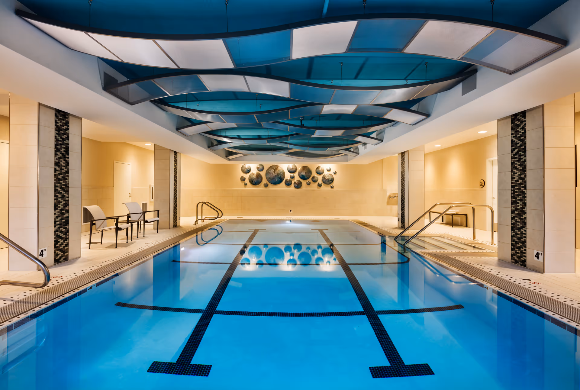 Indoor swimming pool with clear blue water, black lane markings, and a depth marker of 4 feet. The pool area has beige tiled walls and floor, modern blue and white ceiling panels, and decorative circular wall art at the far end. There are metal handrails and a few chairs along the sides.