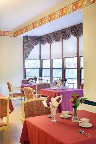 Sunlit dining room with red tablecloths, flowers, and large windows.