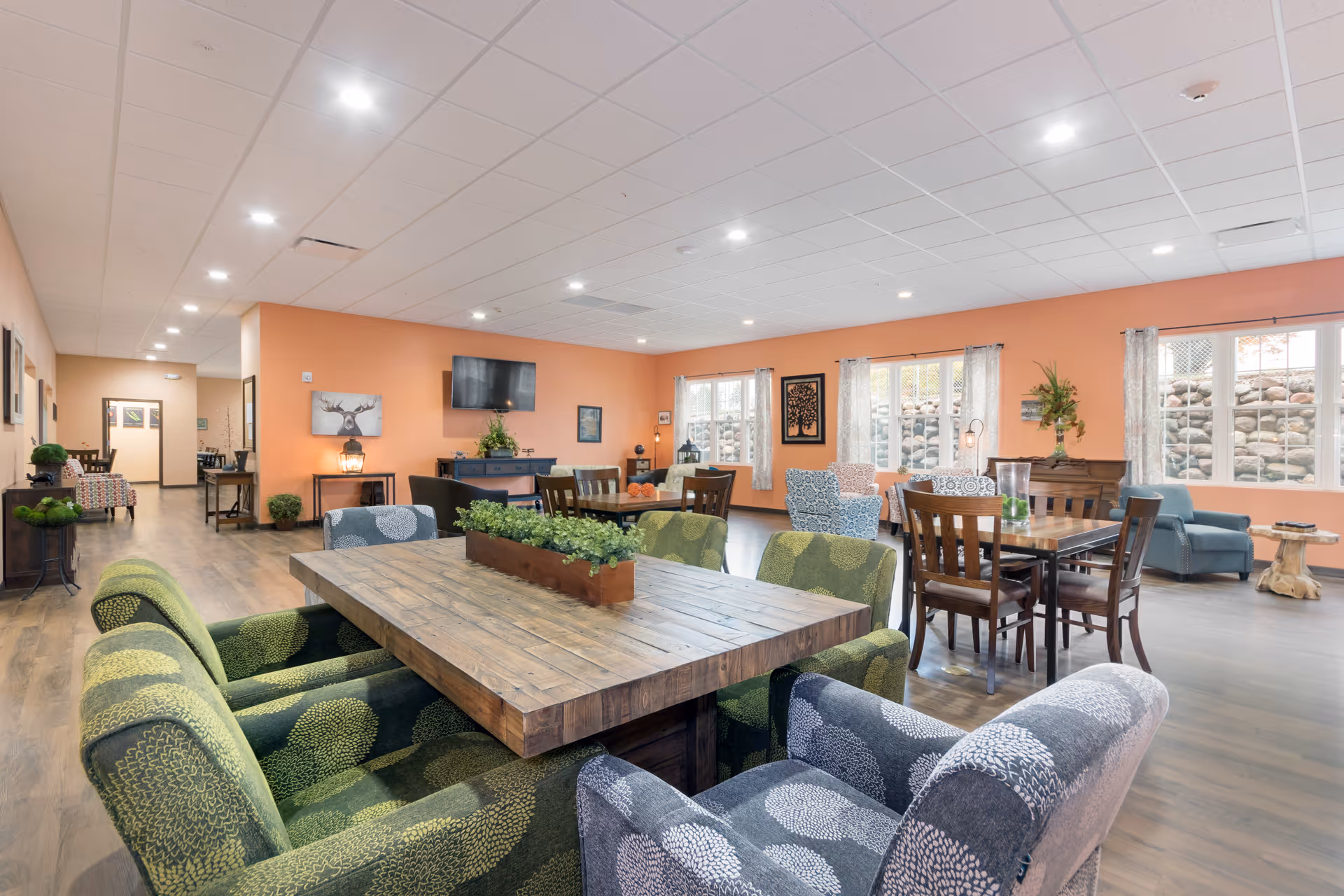 A spacious and well-lit common area in a senior living facility with multiple wooden tables and a variety of upholstered chairs in green and gray patterns. The walls are painted a warm peach color, and large windows with sheer curtains allow natural light to enter. The room features wood flooring, framed artwork, a wall-mounted TV, and decorative plants, creating a comfortable and inviting atmosphere.