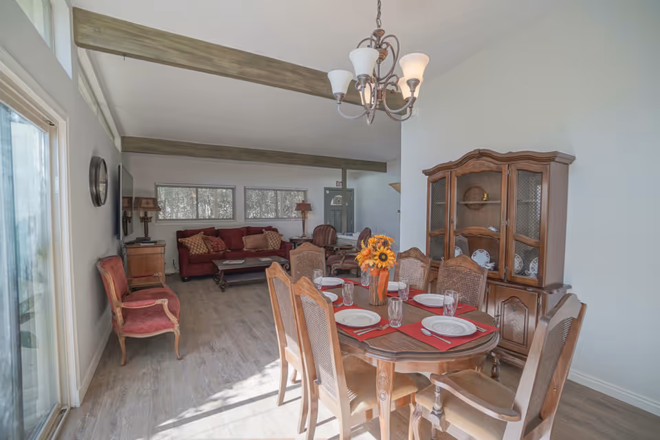 Sunlit dining area with a wooden table set for six and a china cabinet, opening into a living room with red sofas and exposed ceiling beams.