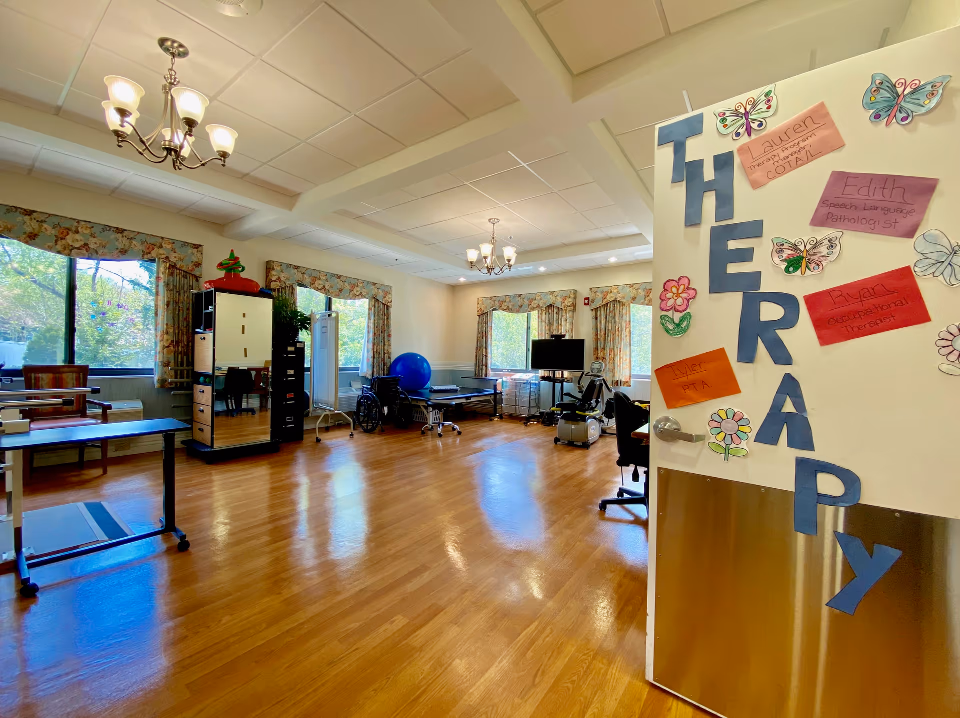Bright therapy/activity room with hardwood floors, exercise equipment, large windows, and a door decorated with 'THERAPY' cutouts.