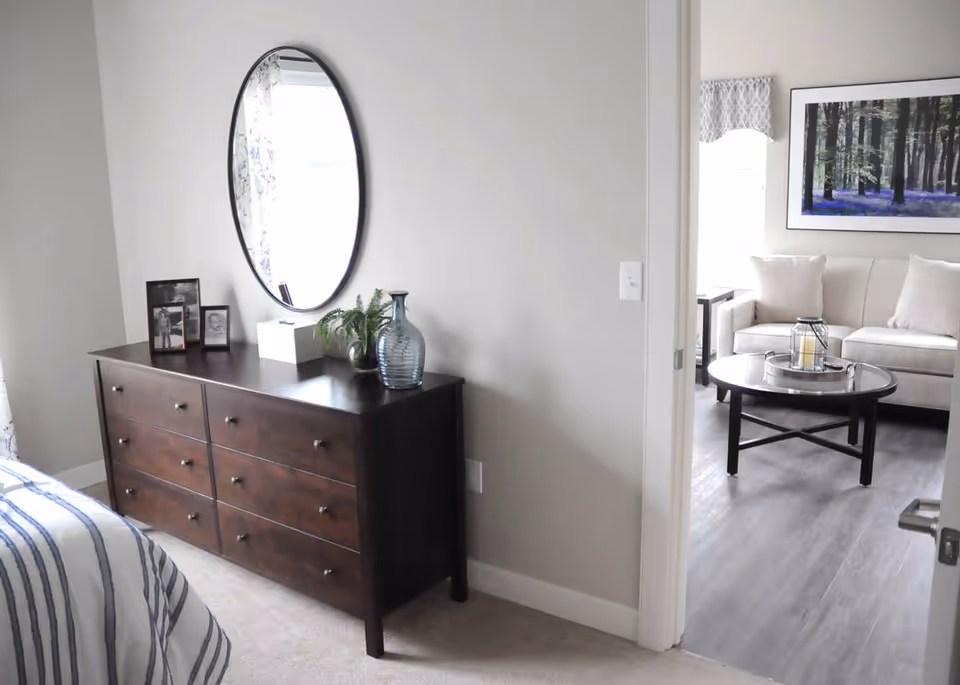 A bedroom corner featuring a dark wooden dresser with six drawers, topped with framed photos, a tissue box, a small plant, and a blue glass vase. Above the dresser is a large oval mirror reflecting light from a window. To the right, an open door leads to a living room with a beige sofa, a round black coffee table with a candle lantern, and a large framed picture of a forest on the wall.