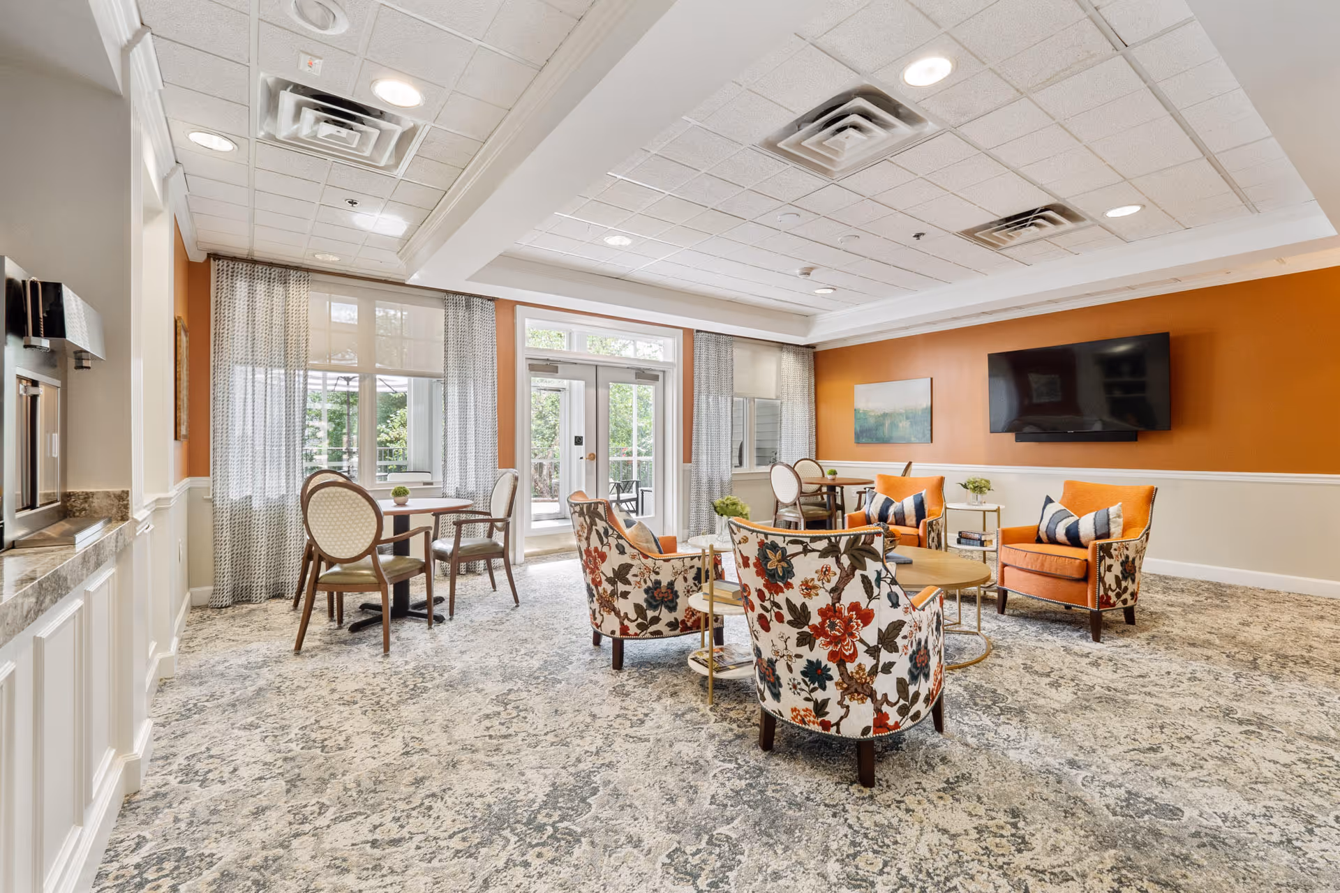 A bright and spacious common area with patterned carpet, floral upholstered chairs, orange armchairs with striped pillows, round wooden tables, and large windows with sheer curtains letting in natural light. A flat-screen TV is mounted on an orange accent wall, and glass doors lead to an outdoor patio.