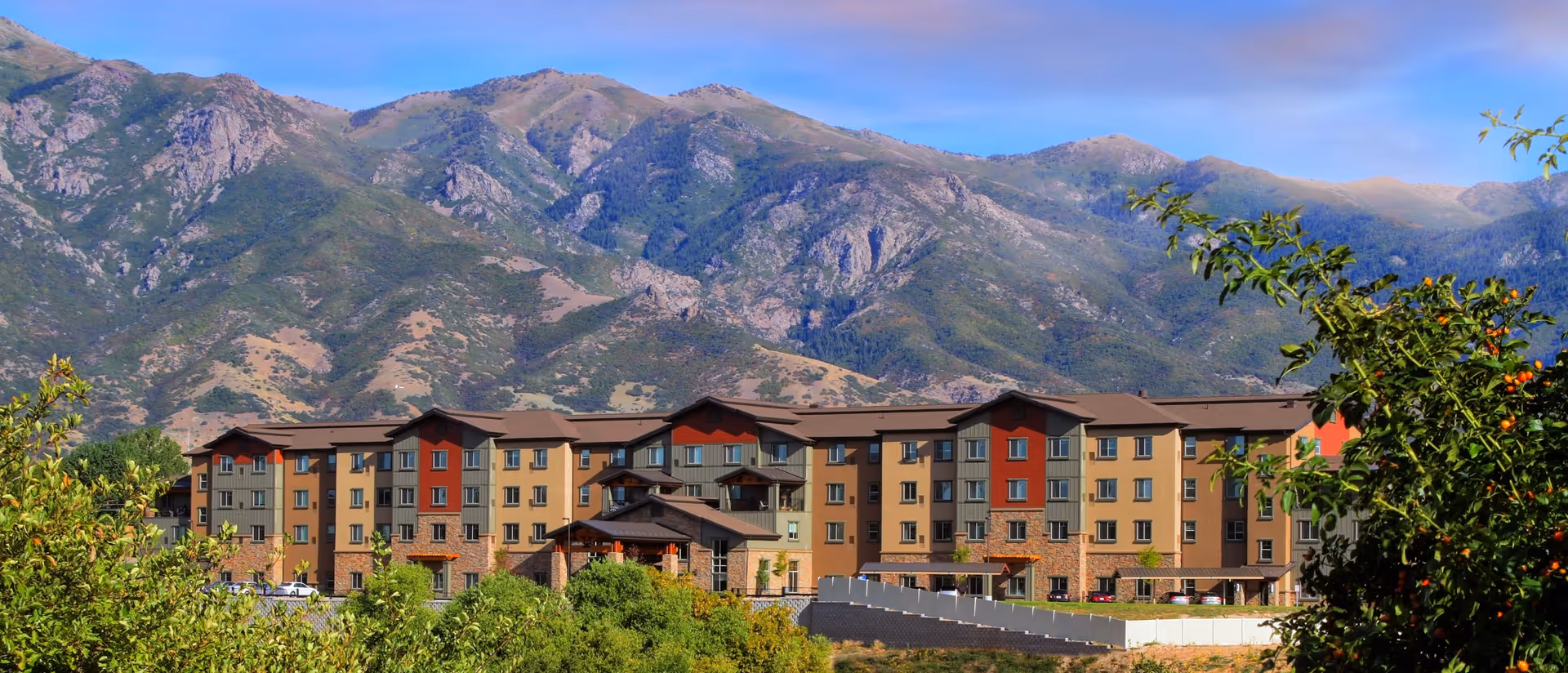 A multi-story senior living community building set against large mountains with trees and shrubs in the foreground.