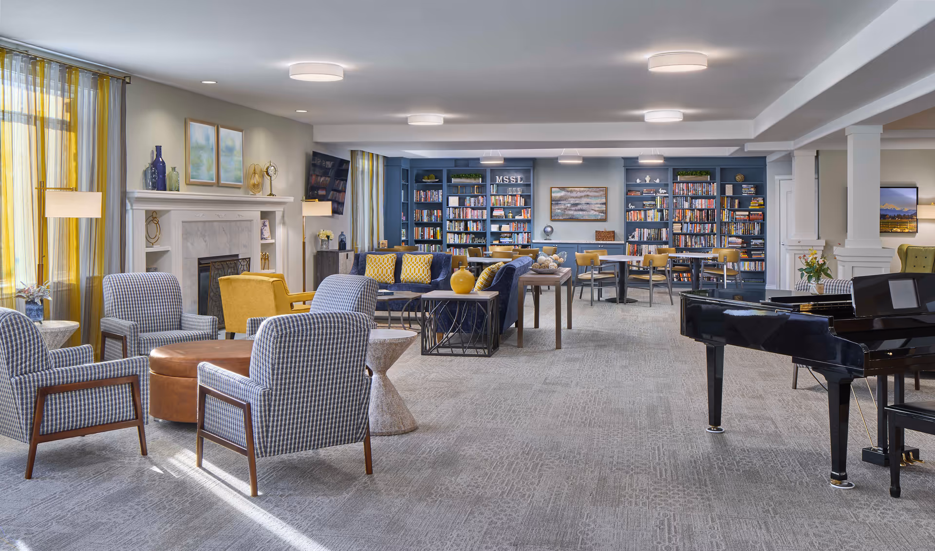 A spacious senior living common area with a variety of seating options including patterned armchairs, yellow armchairs, and blue sofas with yellow pillows. The room features a fireplace with decorative items on the mantle, large windows with yellow and gray curtains, multiple bookshelves filled with books, several tables and chairs, and a black grand piano. The walls are painted light gray and the carpet is a neutral tone.