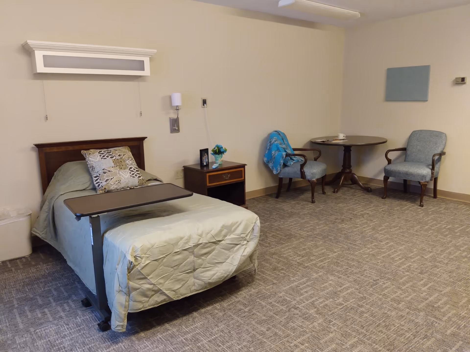 A simple assisted living bedroom with a single bed covered in a light green quilt and a patterned pillow. There is a small bedside table with a flower vase and a framed photo. Two upholstered chairs and a round wooden table with a cup and saucer are placed against the far wall. The room has beige walls and carpeted flooring.