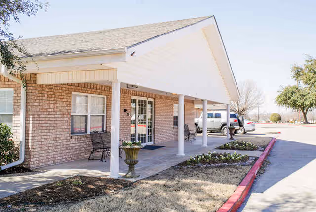 Covered brick front entrance with white columns, benches, a planter, and parked cars along the driveway.
