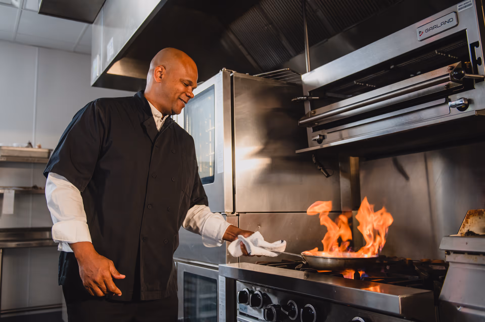 A chef in a commercial kitchen flambés a pan on a gas stove.
