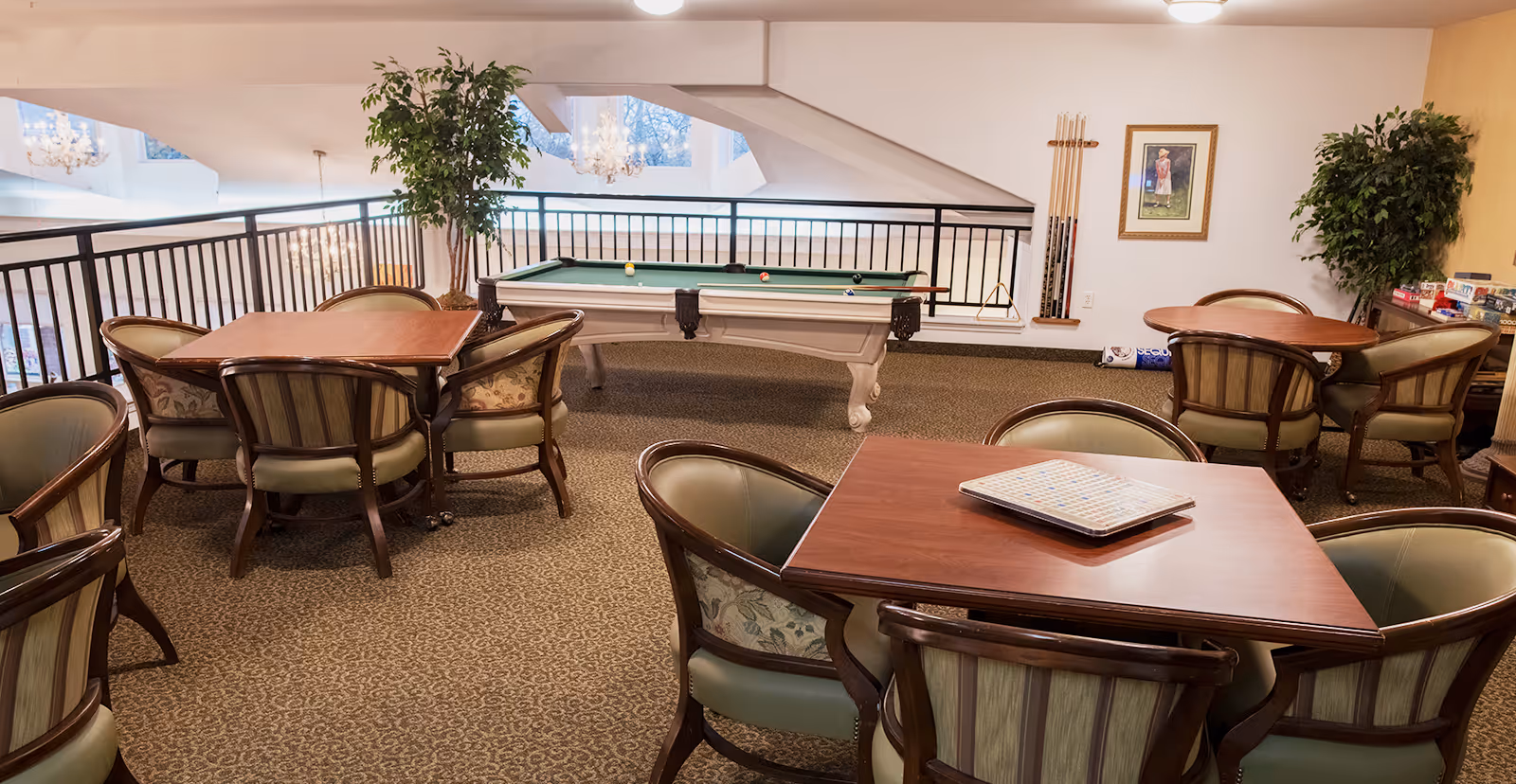 A senior living community game room with several wooden tables and cushioned chairs arranged around them. A pool table with balls and cues is positioned near a railing overlooking a lower level. The room is carpeted and decorated with framed artwork, potted plants, and board games on a side table.