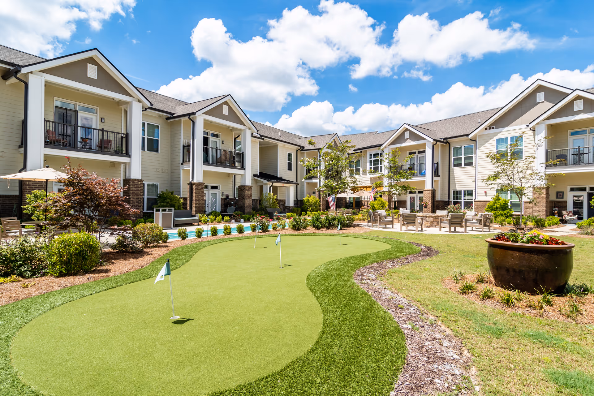 Outdoor courtyard area of Arbor Terrace Hamilton Mill featuring a putting green with small flags, surrounded by well-maintained landscaping, trees, and shrubs. The two-story building with balconies and large windows encloses the courtyard under a bright blue sky with scattered clouds.