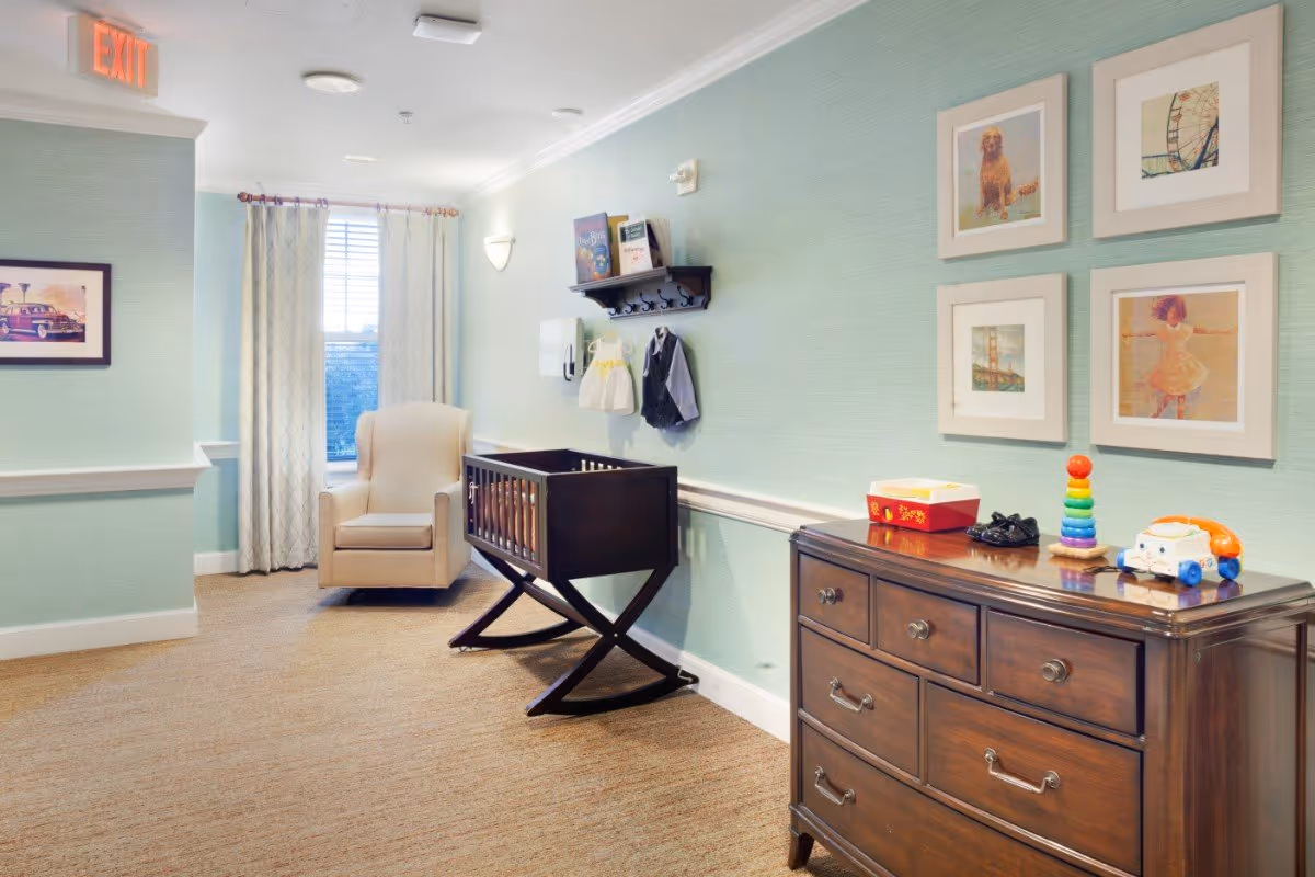 A cozy corner in a senior living facility with a beige armchair next to a window with curtains. A dark wooden baby crib is placed against a light green wall with a shelf holding children's books and two small outfits hanging below it. A dark wooden dresser with toys and framed pictures on the wall above it is also visible. An exit sign is mounted on the ceiling near the corner.