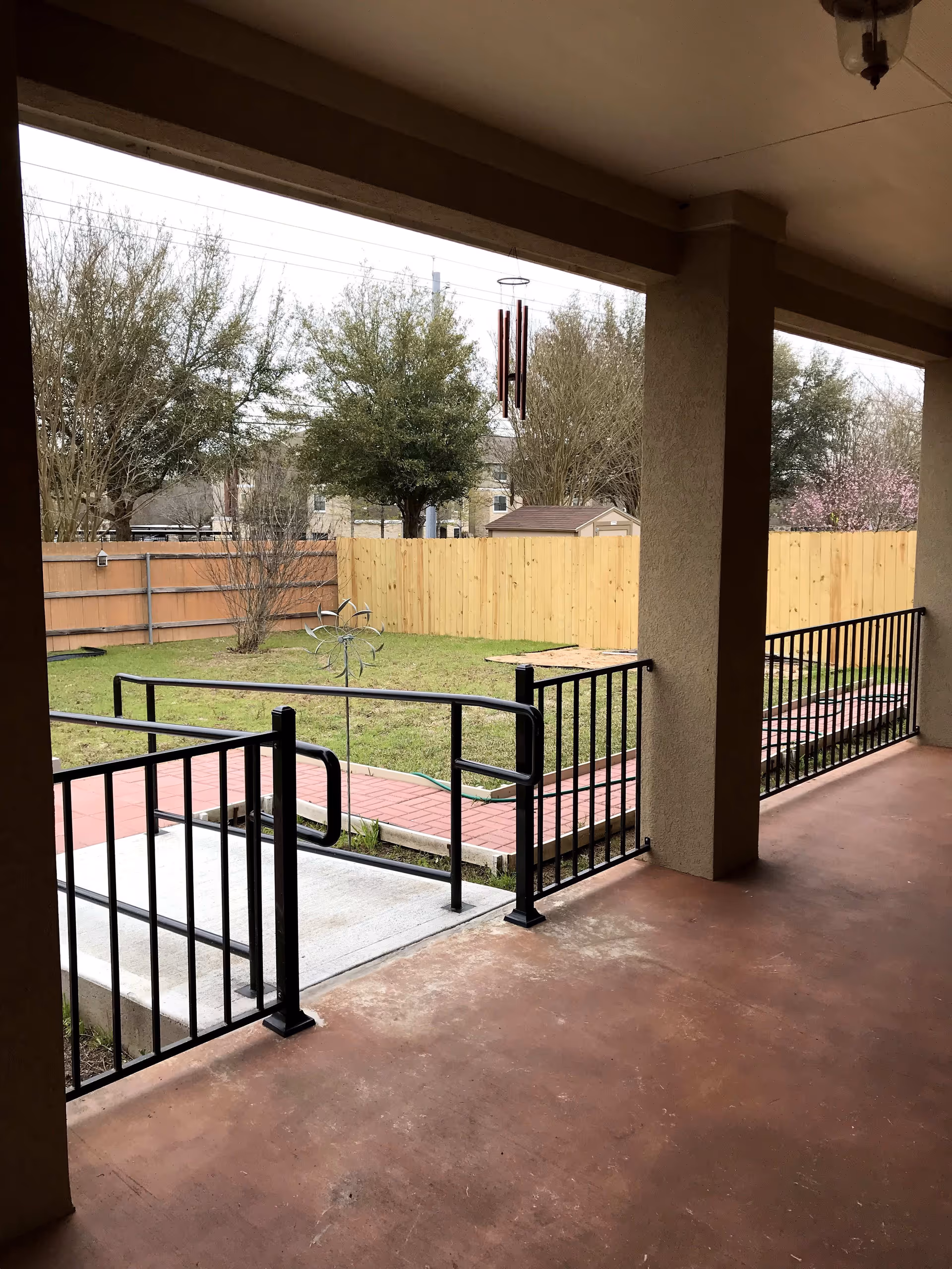 View from a covered patio area with a reddish-brown concrete floor and black metal railings. Beyond the patio is a small grassy yard enclosed by wooden fences, with some trees and a wind spinner decoration. A wind chime hangs from the patio ceiling.