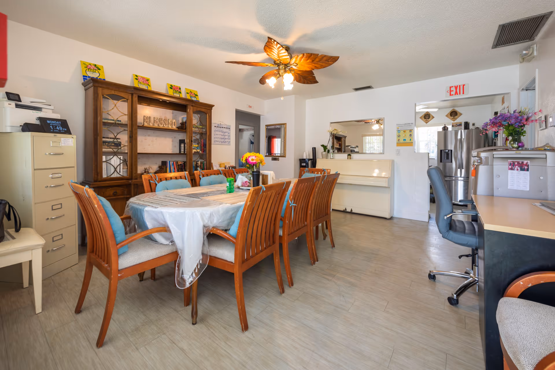 A well-lit room with a long dining table covered with a white tablecloth and surrounded by wooden chairs with blue cushions. There is a wooden cabinet with glass doors and decorative items on top, a white piano against the wall, a ceiling fan with leaf-shaped blades, a filing cabinet, and a desk with an office chair. The room has light-colored flooring and white walls with various decorations and an exit sign above a doorway leading to a kitchen area with a stainless steel refrigerator.