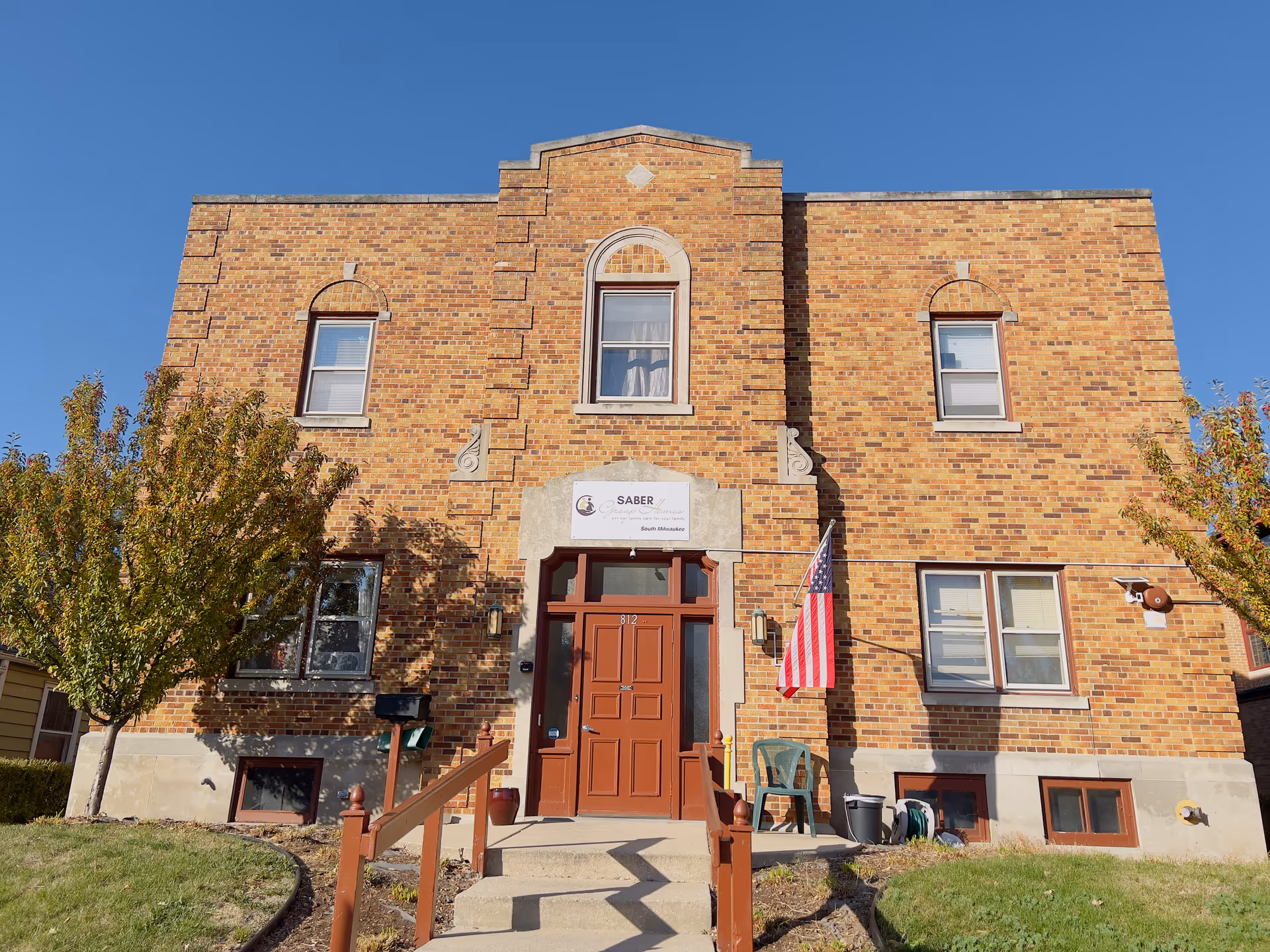 Front exterior view of a two-story brick building with a central entrance door painted brown. There are windows on both floors, an American flag mounted on the right side of the entrance, and two small trees on either side of the walkway leading to the door. A sign above the door reads 'SABER' with additional smaller text below.