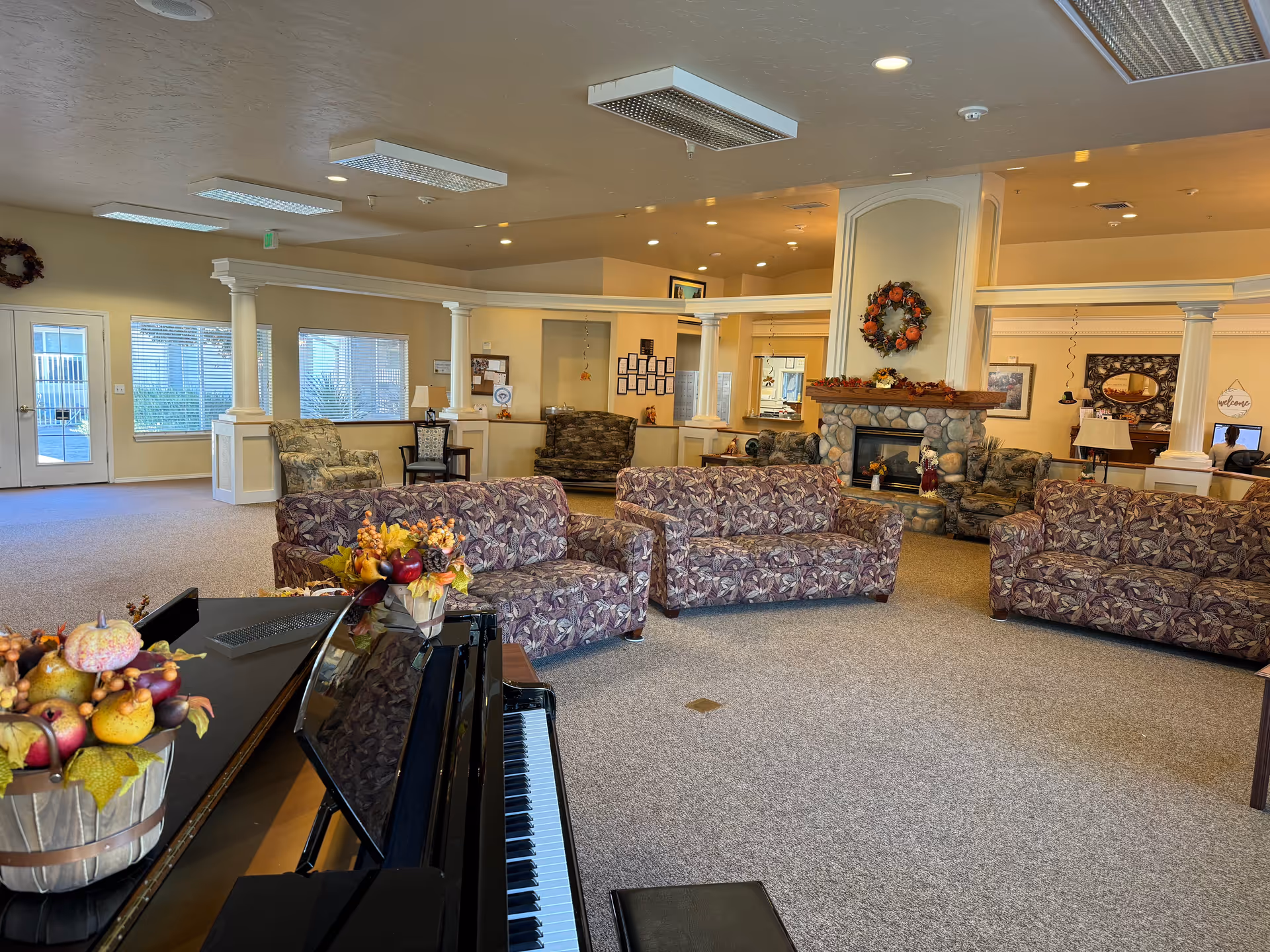 A spacious senior living facility common area with multiple patterned couches arranged around a stone fireplace decorated with a fall wreath and seasonal decor. A black piano with a fall-themed floral arrangement is in the foreground. The room has beige walls, carpeted floors, white columns, and large windows letting in natural light.