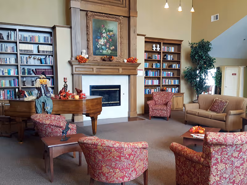 Cozy assisted-living common room with a fireplace flanked by bookshelves, a grand piano and upholstered chairs and sofa.