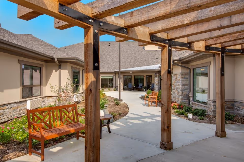 Outdoor courtyard area at The Shores at Clear Lake featuring wooden benches, a small table with a plant, a pergola structure overhead, and surrounding garden beds with flowers and shrubs. The building exterior has beige walls with stone accents and multiple windows.