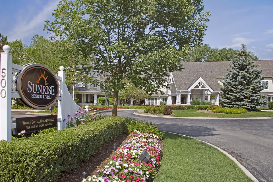 Front entrance of Sunrise Senior Living with a large sign, circular driveway, manicured lawns, and flowerbeds in front of the building.