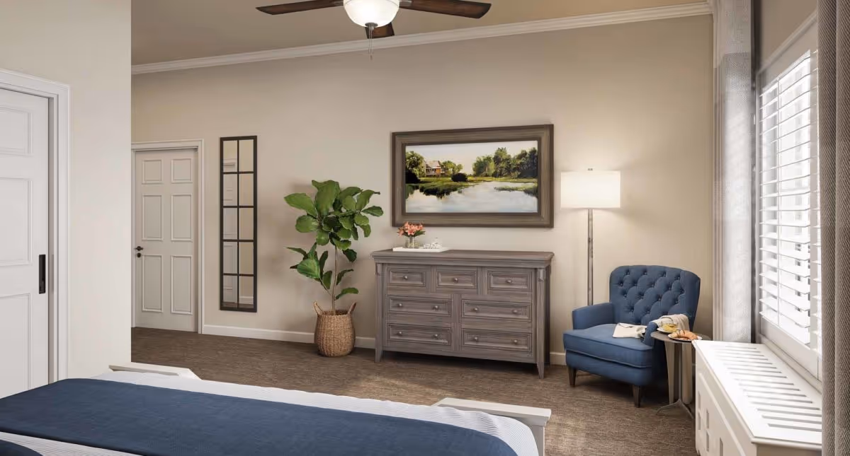 A cozy bedroom interior featuring a bed with a blue blanket in the foreground, a gray wooden dresser against the wall with a framed landscape painting above it, a tall green potted plant to the left, a blue upholstered armchair with a small side table holding a plate of food and a drink, a floor lamp beside the chair, and windows with white shutters on the right side.