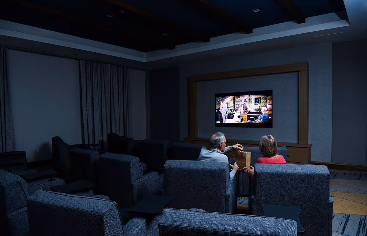 A dimly lit movie theater room with multiple rows of gray upholstered seats. Two elderly people, a man and a woman, are seated in the front row watching a movie on a wall-mounted flat screen TV. The man is holding a bag of popcorn.