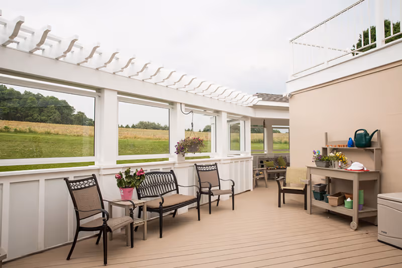 A covered outdoor patio area with beige flooring and white railings. The space features several chairs and a bench with cushions, a small table with a potted plant, and a gardening station with pots, a watering can, and gardening tools. Beyond the patio, there is a grassy field and trees under a cloudy sky.