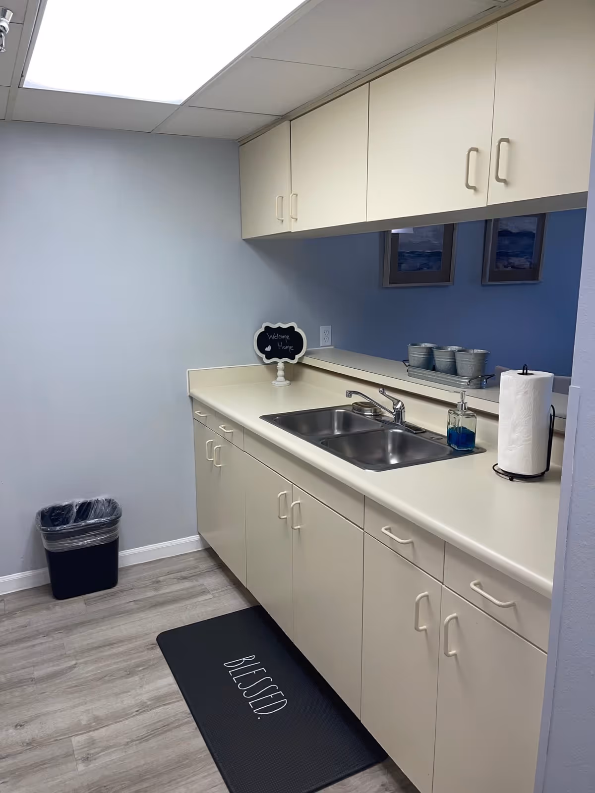 Small kitchenette with white cabinets, a stainless double sink, paper towel holder, soap dispenser, trash can, and a black mat reading 'BLESSED'.