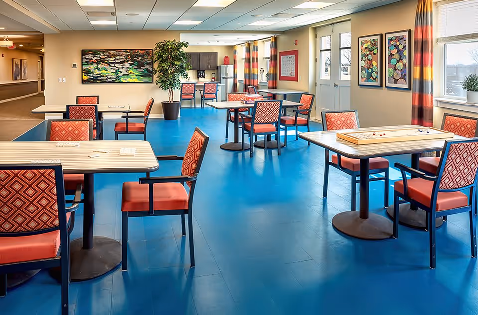 A bright and spacious common area with blue flooring, several tables and chairs with red patterned upholstery, colorful curtains, and framed artwork on the walls. There is a kitchenette area with a coffee maker and refrigerator in the background, and a hallway leading to other rooms.