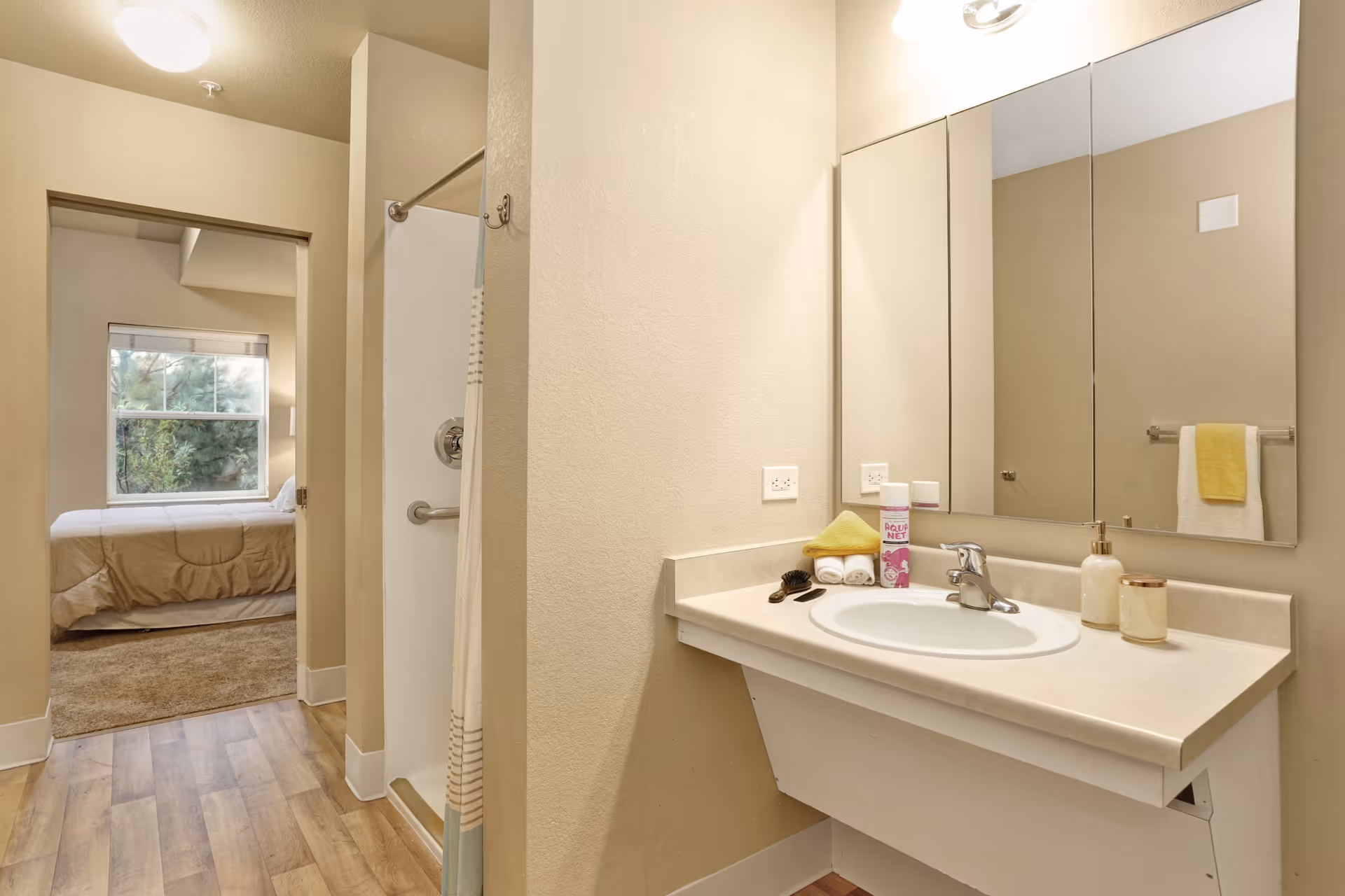 Interior view of a bathroom with a sink, mirror, and toiletries on the counter. To the left, there is a shower with a curtain and a doorway leading to a bedroom with a bed and a window showing greenery outside.