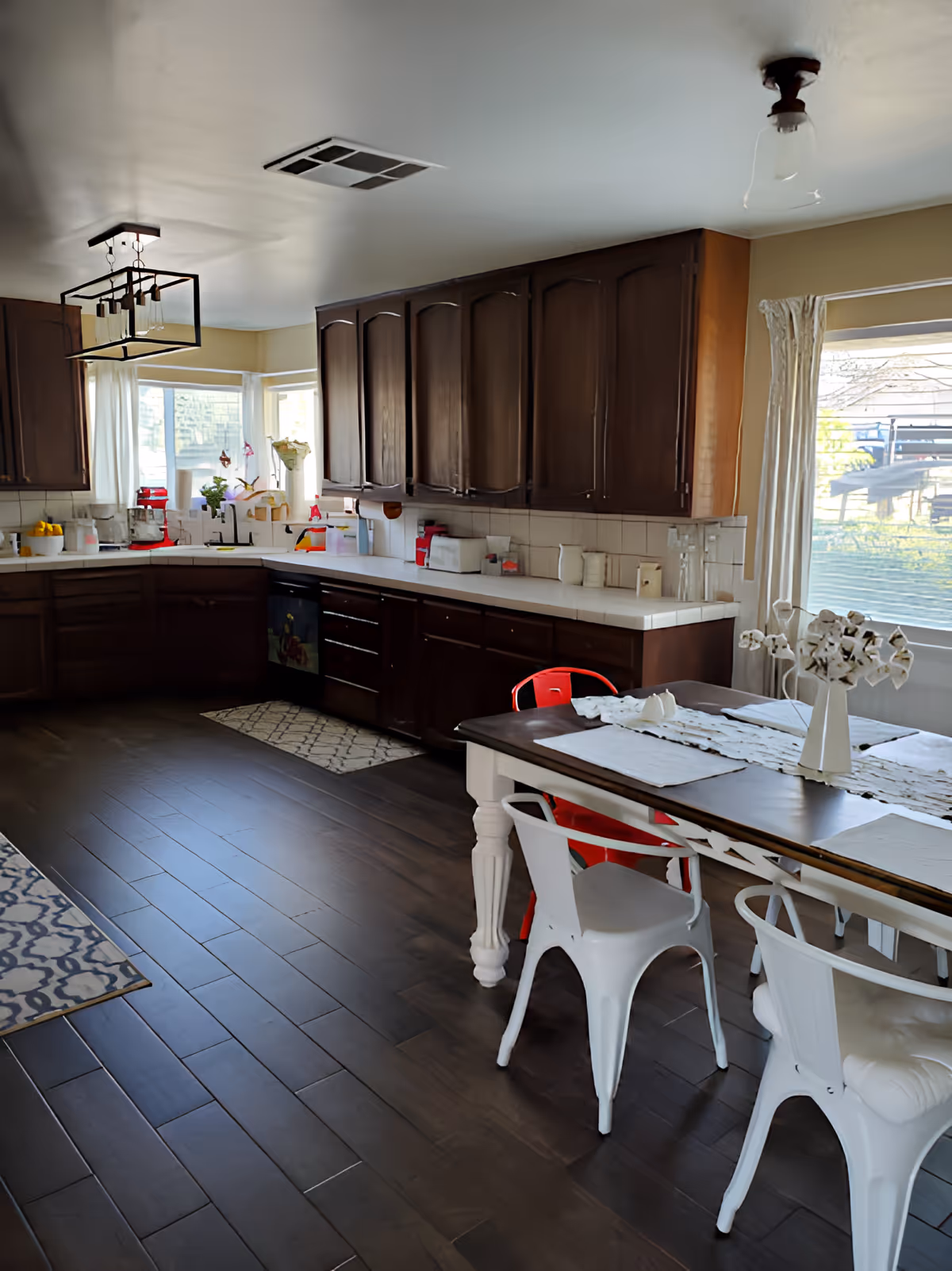 A spacious kitchen with dark wooden cabinets, white countertops, and a dark wooden floor. There is a dining table with white and red metal chairs, a vase with flowers on the table, and large windows with white curtains letting in natural light.