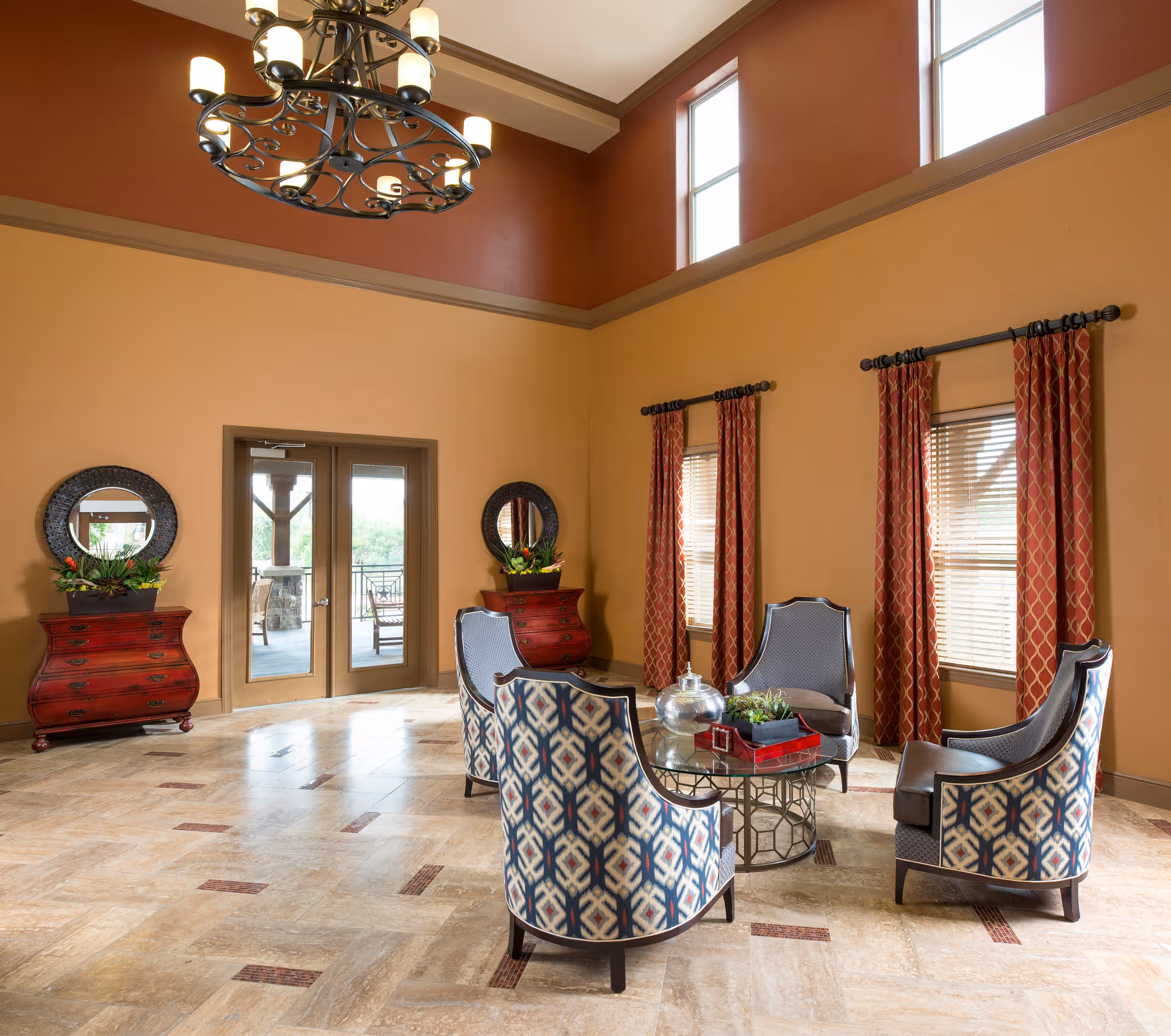 A spacious and well-lit living room area with high ceilings and large windows. The room features four patterned armchairs arranged around a glass coffee table with decorative items on top. There are two red wooden dressers with round mirrors and plants on either side of a double glass door leading outside. The walls are painted in warm tones of beige and terracotta, and the floor is tiled with a light stone pattern. A large, ornate chandelier hangs from the ceiling.