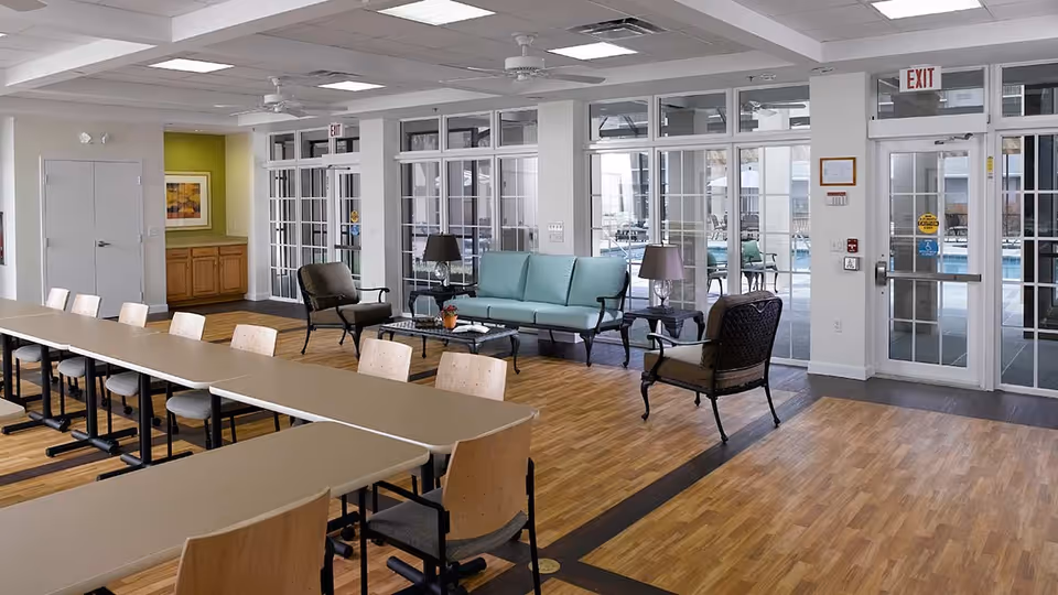 A spacious interior room with a long table and multiple chairs arranged for meetings or dining. There is a seating area with a light blue sofa, two armchairs, and two side tables with lamps. Large windows and glass doors provide a view of an outdoor pool area. The room has wood flooring and a ceiling with recessed lighting and ceiling fans.