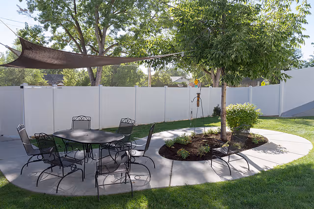 Outdoor patio area with a round metal table and six matching chairs on a concrete surface. A shade sail is stretched overhead, and there is a curved walkway around a small landscaped area with a tree and plants. A white fence encloses the space, and green grass surrounds the patio.