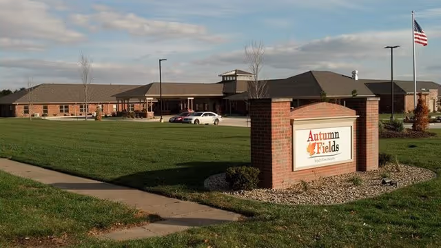 Exterior view of Autumn Fields Adult Community-Hoopeston building with a large green lawn, a sidewalk, a brick sign displaying the facility name, several parked cars, and an American flag on a flagpole under a partly cloudy sky.