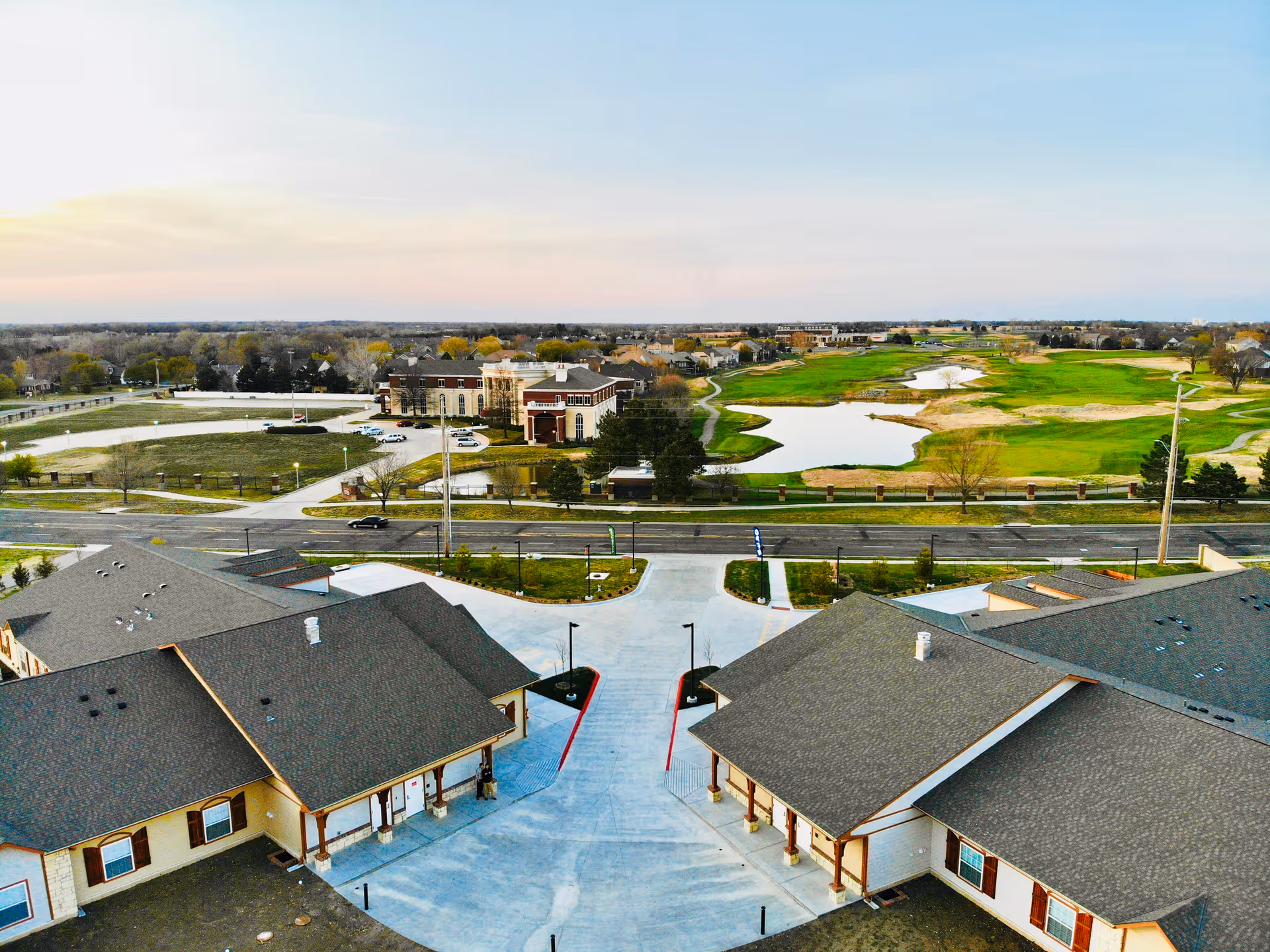 Aerial view of The Mapleton Assisted Living facility showing two large buildings with gray roofs and beige walls separated by a wide concrete driveway. Across the street, there is a large building with a parking lot and a green golf course with water features extending into the distance under a clear sky.